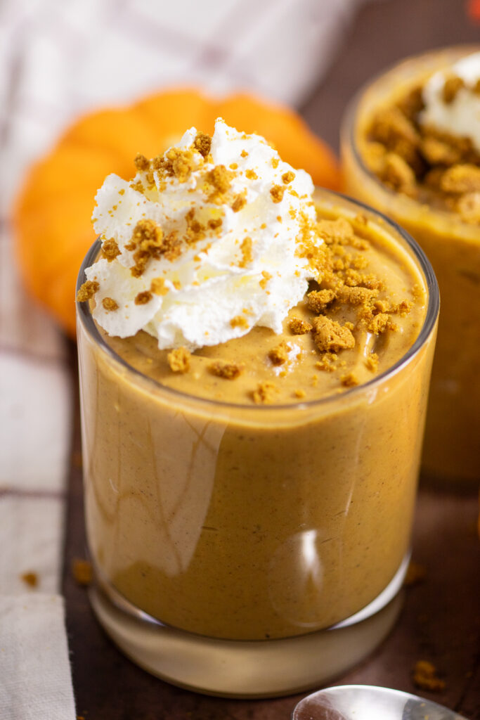 Pumpkin pudding in a small glass topped with whipped cream and gingersnap crumbs with another glass of pudding behind. They're surrounded by mini pumpkins and a linen napkins.