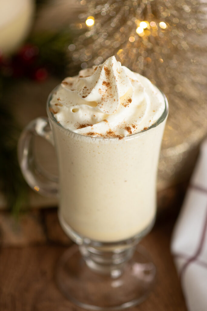 An overhead view of a glass mug of homemade eggnog topped with a swirl of whipped cream and a sprinkle of cinnamon on a rustic wood background next to a linen napkin.
