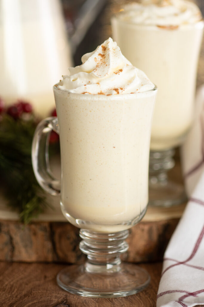 A glass mug of homemade eggnog topped with whipped cream on a rustic wood background with Christmas greenery and another mug of eggnog behind.