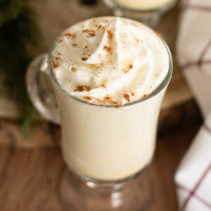 An overhead view of a glass mug of homemade eggnog topped with a swirl of whipped cream and a sprinkle of cinnamon on a rustic wood background next to a linen napkin.