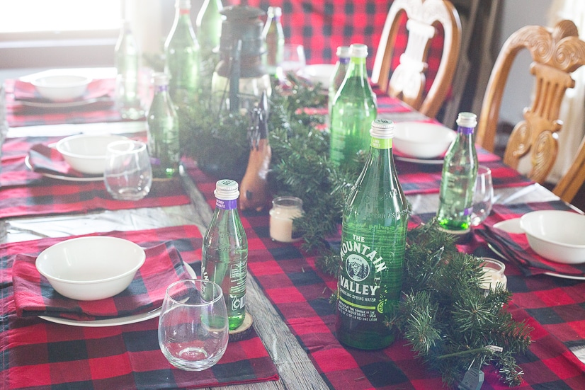Rustic Christmas and Holiday table decorations and table settings with a lantern centerpiece, evergreen garland, wood slice coasters, red and black buffalo check, and green Mountain Valley Spring Water bottles.