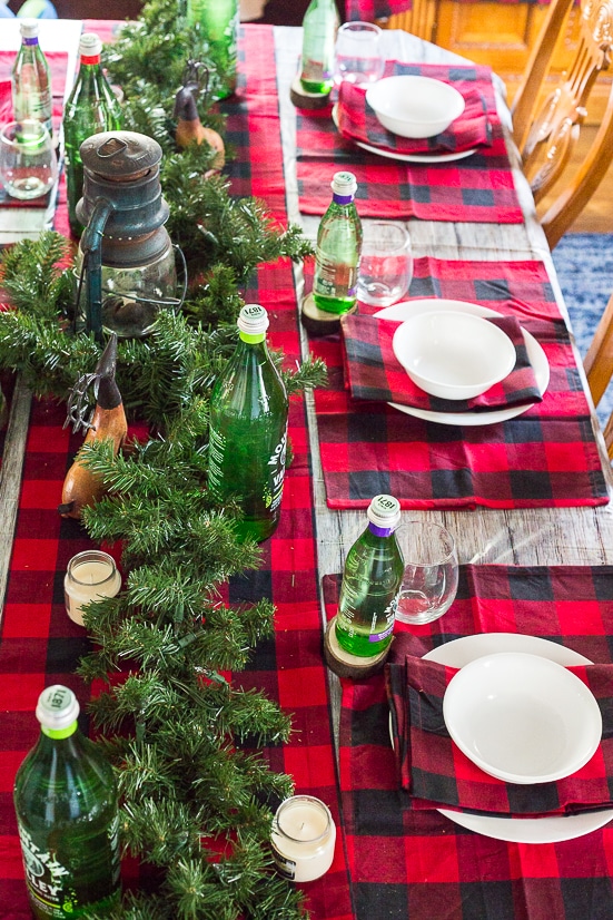 Rustic Christmas and Holiday table decorations and table settings with a lantern centerpiece, evergreen garland, wood slice coasters, red and black buffalo check, and green Mountain Valley Spring Water bottles.