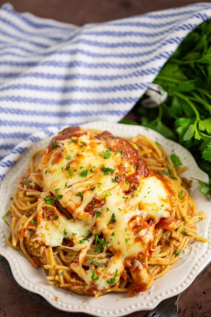 Overhead view of one dish cheesy chicken pasta bake on a small white plate topped with melted mozzarella and freshly chopped parsley on a rustic wood background.