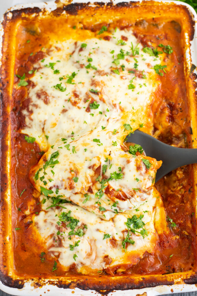 Overhead view of One Dish Cheesy Chicken pasta bake, showing the chicken breast with melted mozzarella and freshly chopped parsley with a spoon under one of the chicken breasts.