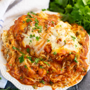 Chicken breast topped with red pasta sauce and melted mozzarella on a small plate filled with baked spaghetti with a linen napkin and bunch of parsley behind.