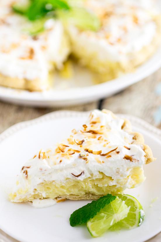 Slice of coconut cream pie on a small plate topped with toasted coconut next to two lime wedges