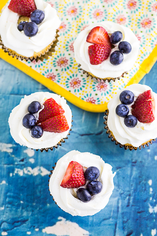 Overhead shot of cupcakes with white frosting topped with strawberries and blueberries on a blue concrete background