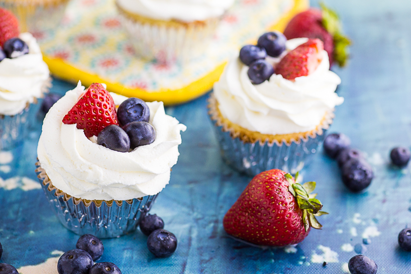 Cupcake topped with strawberry and blueberries on a blue concrete background with more cupcakes in the background
