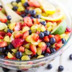 Large glass bowl of fruit salad with grapes, strawberries, and blueberries topped with lemon and lime wedges with a wooden spoon in the middle.