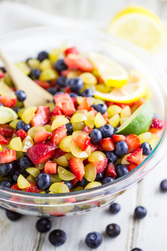 Large glass bowl of fruit salad with grapes, strawberries, and blueberries topped with lemon and lime wedges with a wooden spoon in the middle.