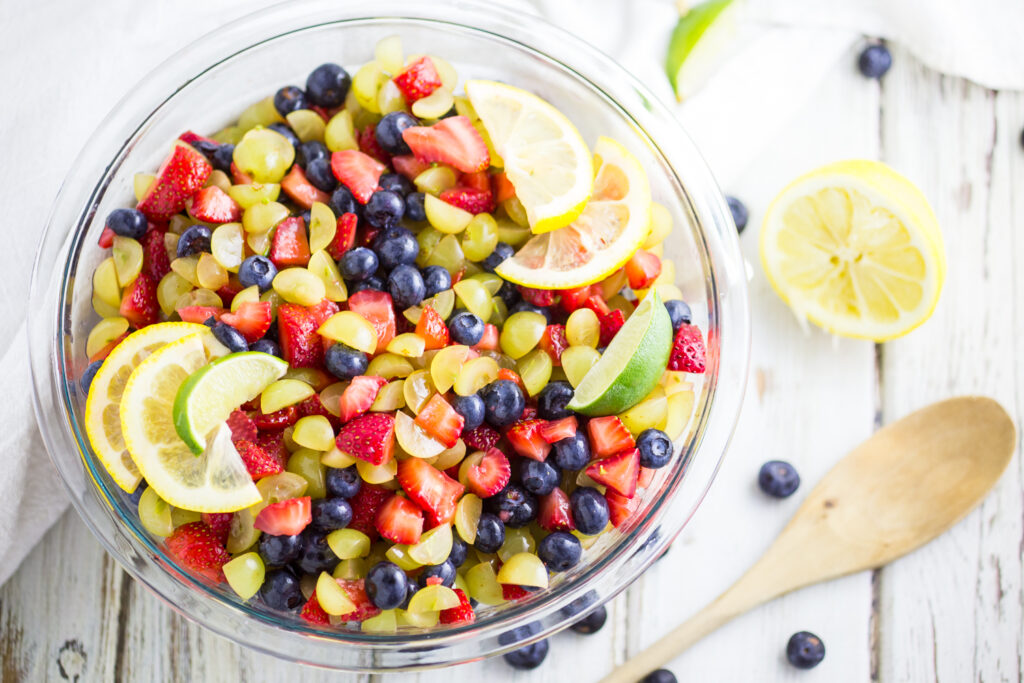 Overhead view of homemade fruit salad topped with fresh lemon and lime slices on a white wooden background next to a wooden spoon.