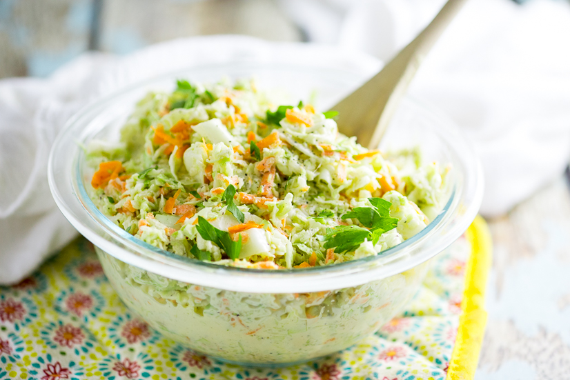 Creamy coleslaw in a clear glass bowl on a yellow flower pot holder with a wooden spoon sticking out