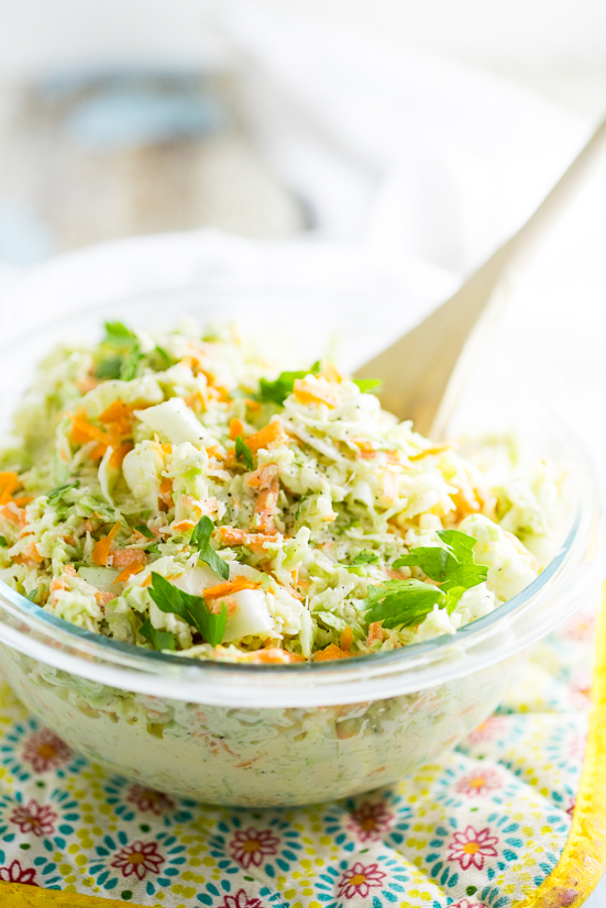 Creamy coleslaw in a clear glass bowl on a yellow flower pot holder with a wooden spoon sticking out