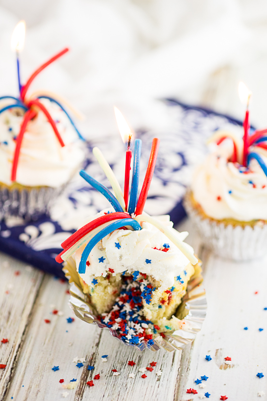 Vanilla cupcake with red, white, and blue star sprinkles, topped with a pull 'n peel twizzler and a sparkler candle on a rustic white wood background. 