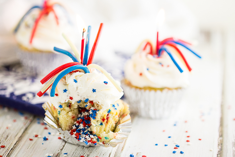 Vanilla cupcake with red, white, and blue star sprinkles, topped with a pull 'n peel twizzler and a sparkler candle on a rustic white wood background.