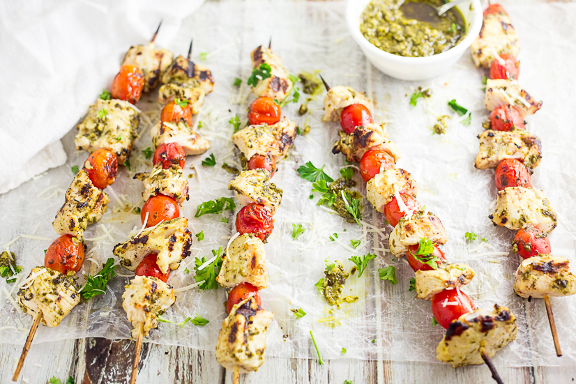 Grilled chicken and cherry tomatoes on skewers brushed with pesto sitting on wax paper with a small white bowl of pesto on a white distressed wood background