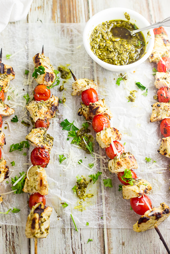 Grilled chicken and cherry tomatoes on skewers brushed with pesto sitting on wax paper with a small white bowl of pesto on a white distressed wood background