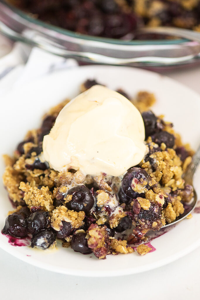 Blueberry crisp topped with vanilla ice cream on a small white plate with a white marble background