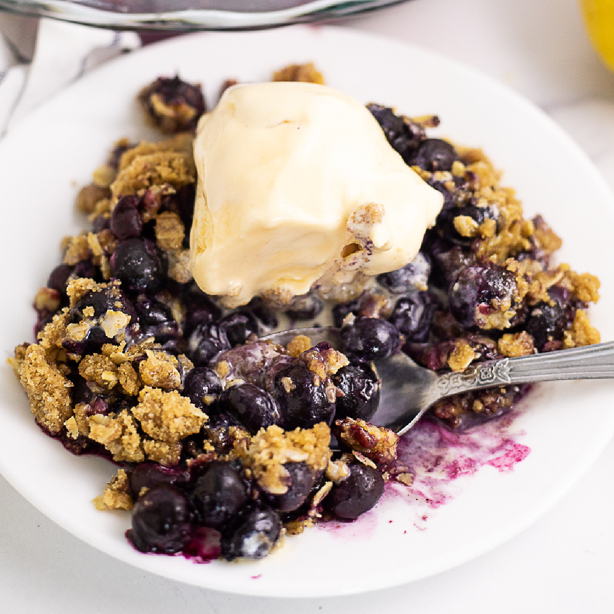 Blueberry crisp topped with vanilla ice cream with a spoon in the middle on a small white plate with a white marble background
