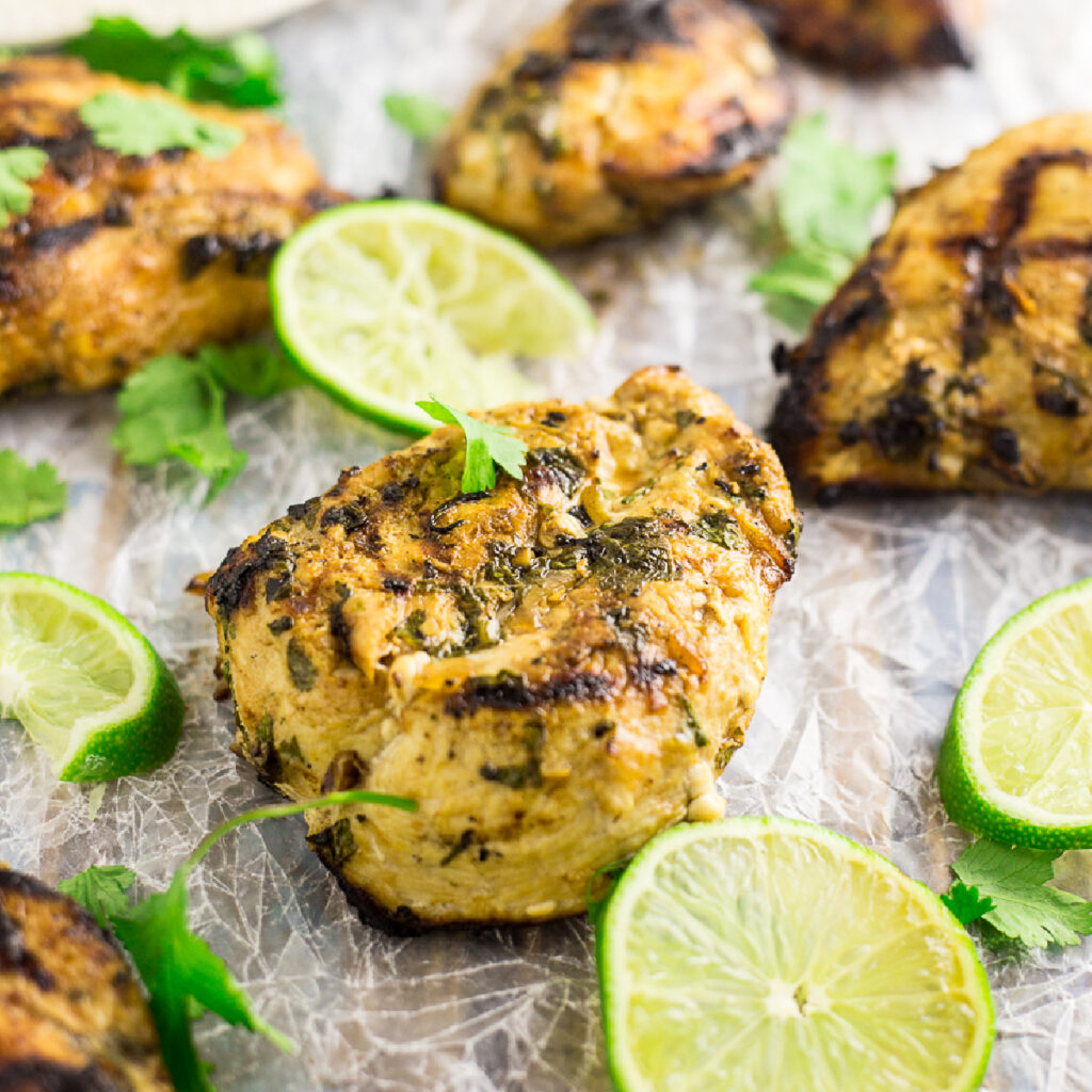 Grilled Cilantro Lime Chicken thigh surrounded by lime slices, fresh cilantro leaves, and more chicken on a piece of parchment paper