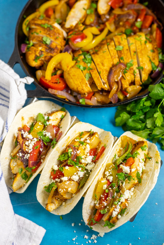 Overhead view of three chicken fajitas in flour tortillas on a bright blue background next to a linen napkin, a fresh bunch of cilantro, and a cast iron skillet with more sauteed peppers and onion and sliced chicken.