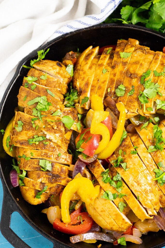 Overhead view of chicken fajitas, made with chicken breast, bell pepper, and onion, in a large cast iron skillet topped with fresh cilantro, next to a white linen napkin.