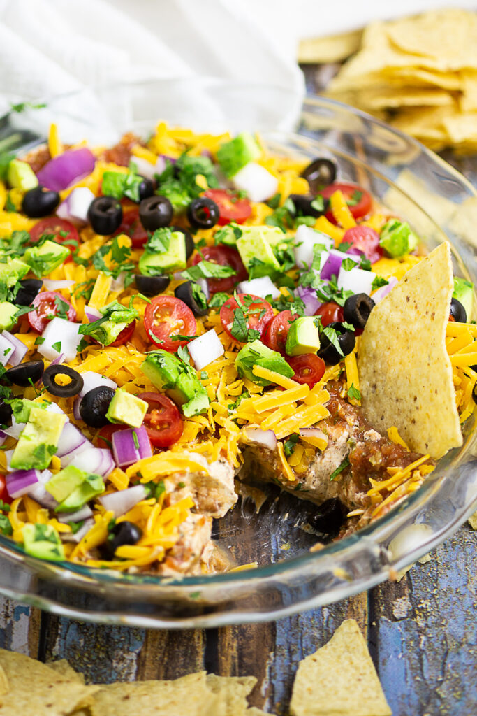 Layered taco dip in a round glass dish with a scoop missing and a chip sticking out of it. Tortilla chips and clean linen surround the dish