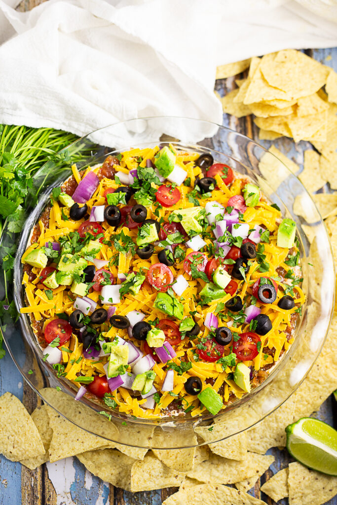 Overhead view of easy layered taco dip in a round glass dish surrounded by tortilla chips, a fresh bunch of cilantro, lime wedges and a clean linen.