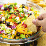 Easy layered taco dip in a round glass dish with a tortilla chip dipping into it
