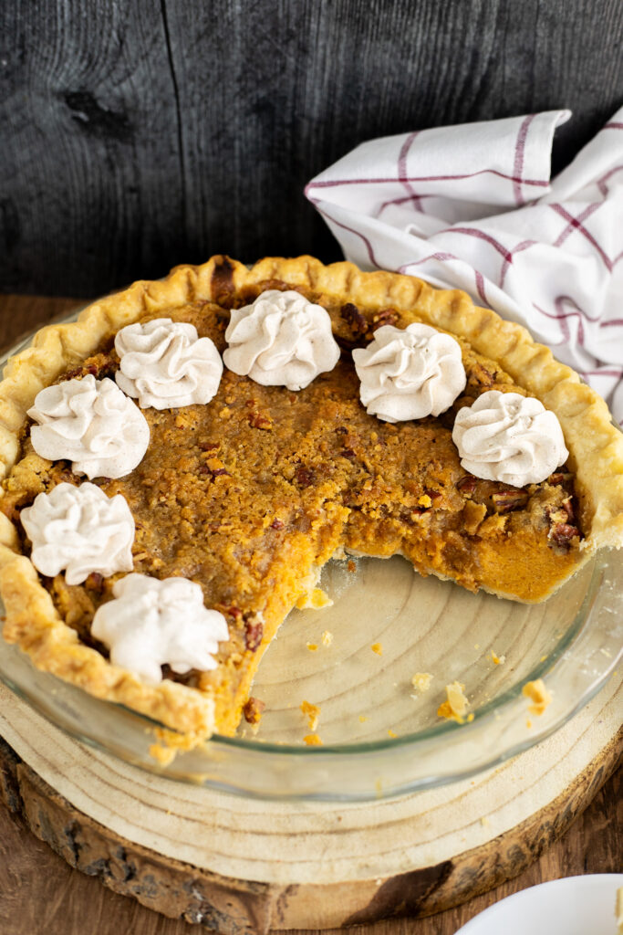 A southern sweet potato pie in a glass pie dish topped with swirls of whipped cream, all on a rustic wood background with a linen napkin behind.