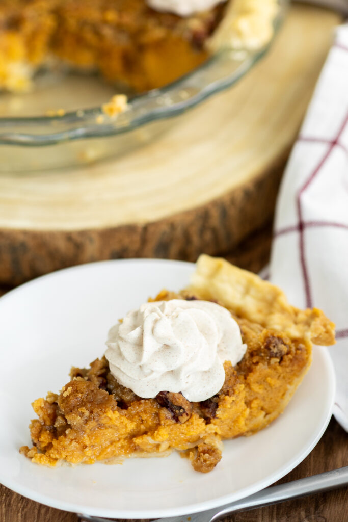 A piece of southern sweet potato pie with a brown sugar pecan crumble on top and a swirl of whipped cream sitting on a small plate with the remaining pie sitting behind it.