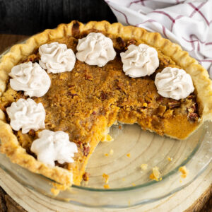 A southern sweet potato pie in a glass pie dish topped with swirls of whipped cream, all on a rustic wood background with a linen napkin behind.