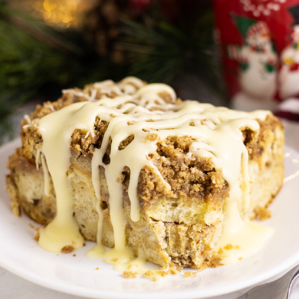 A piece of eggnog French Toast casserole on a small plate topped with eggnog glaze with a red mug and Christmas lights behind.
