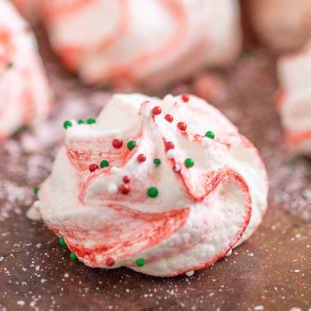 Red and white striped Peppermint Meringue cookie topped with red and green nonpareils on a baking sheet background surrounded by crushed peppermint candy dust.