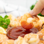 Shrimp dipping into a small glass bowl of homemade shrimp cocktail sauce surrounded by more shrimp.