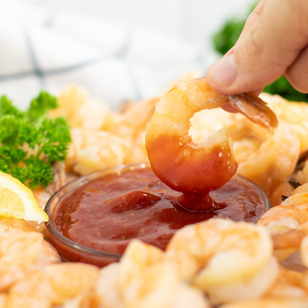 Shrimp dipping into a small glass bowl of homemade shrimp cocktail sauce surrounded by more shrimp.