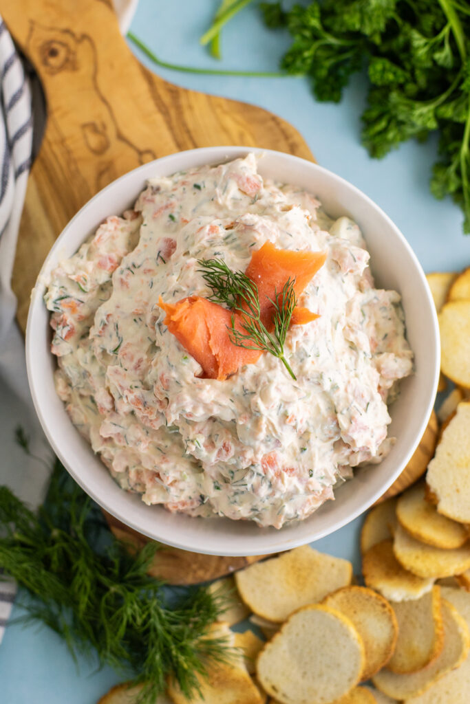 Overhead view of smoked salmon dip in a bowl garnished with a slice of smoked salmon and a sprig of fresh dill. The bowl is sitting on a small cutting board next to a pile of bagel chips, fresh dill, and fresh parsley.