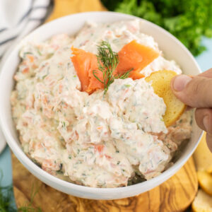 Smoked Salmon Dip in a white ceramic bowl topped with slices of smoked salmon and a sprig of fresh dill with a bagel chip dipping in.