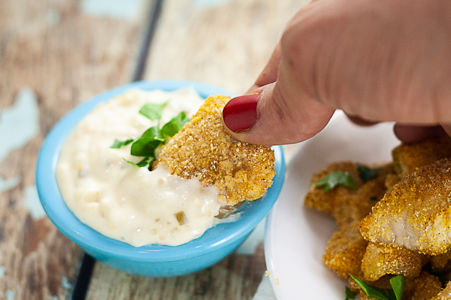 Cornmeal coated baked catfish nugget being dipped into a small blue bowl filled with tartar sauce on a rustic blue wood background