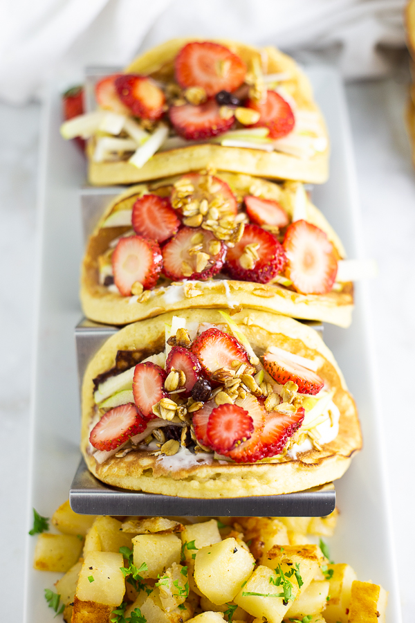 Pancake parfait tacos in a metal taco stand on a rectangle white plate with seasoned breakfast potatoes