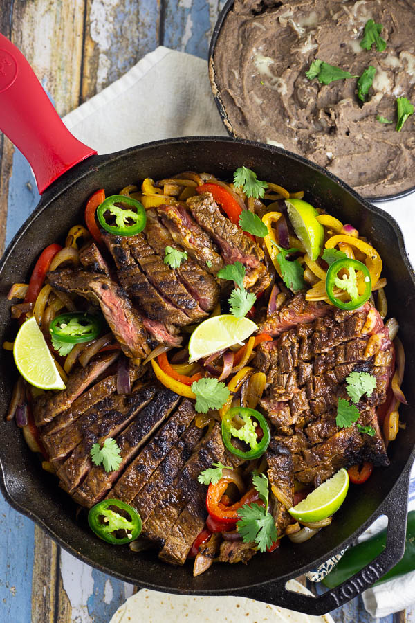 Steak fajitas in  a black cast iron skillet with red and yellow bell peppers and fresh slices of jalapenos and lime.