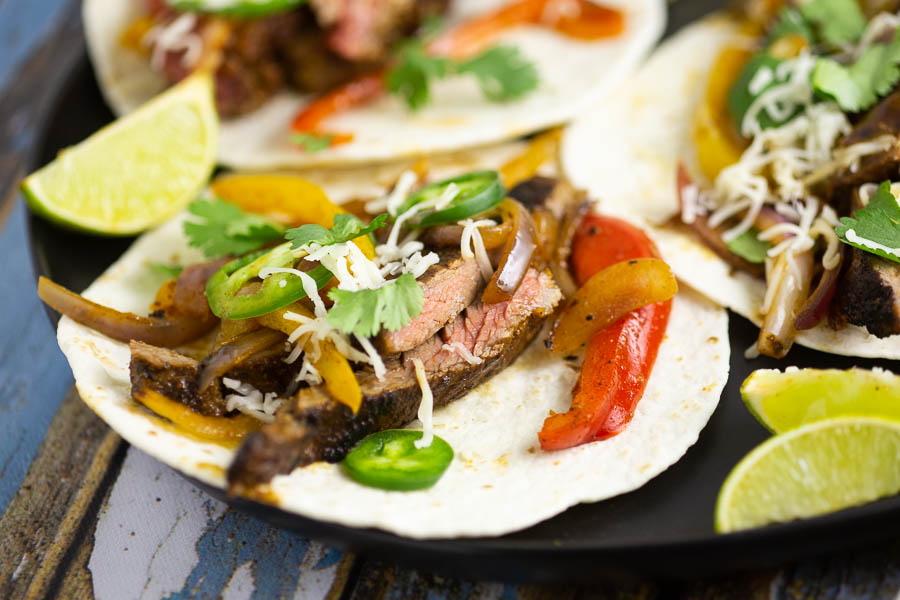 Close up of a steak fajita on a flour tortilla sitting on a black place with lime wedges