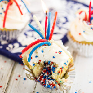 Vanilla cupcake filled with red, white, and blue star sprinkles topped with a peeled licorice and a red candle with more cupcakes in the background