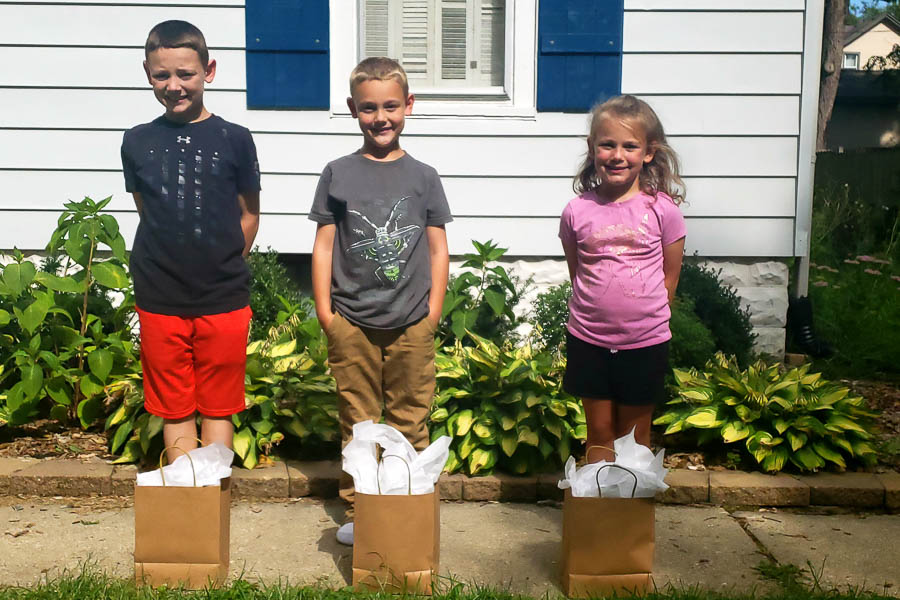 3 kids standing in front of a white house with brown bags