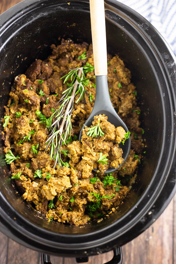 Large oval slow cooker crock filled with crockpot stuffing topped with fresh parsley and a sprig of rosemary with a wooden and silicone spoon in the middle on a rustic wood background.