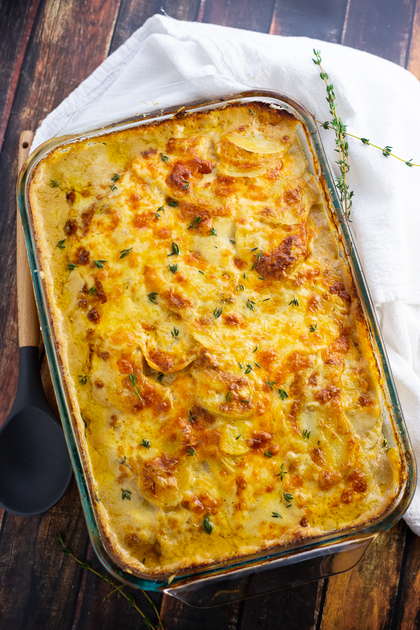 Casserole dish filled with scalloped potatoes with fresh thyme on top, on top of a fresh white linen on a rustic background