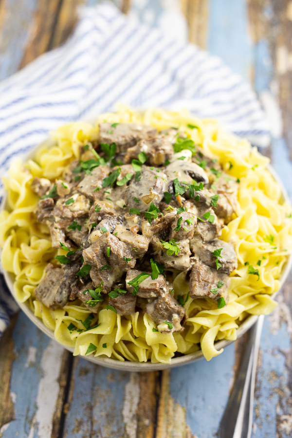Slow cooker beef stroganoff over egg noodles topped with parsley in a white bowl on a blue distressed wood background and a white and blue striped linen