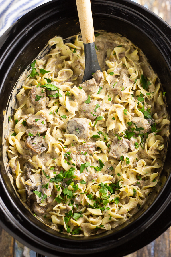 Slow cooker beef stroganoff in a black Crock Pot with a wooden spoon sticking out