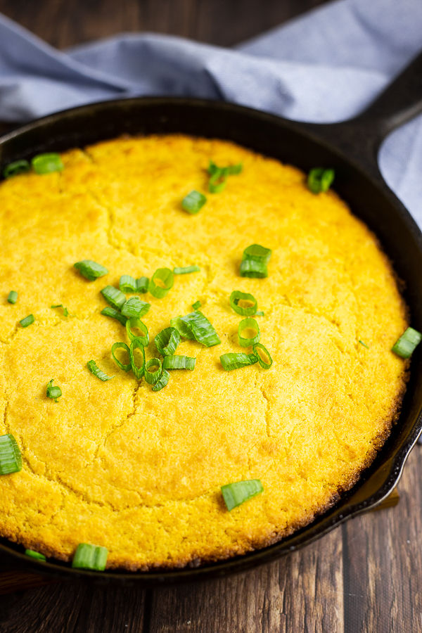 Southern skillet cornbread topped with scallions in a black cast iron skillet with a blue canvas linen on a rustic wood background.