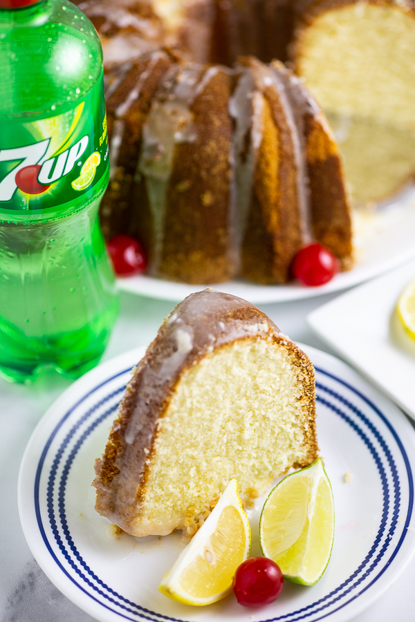 A slice of 7Up pound cake on a blue and white striped plate with a lemon wedge, lime wedge, and cherry, and a bottle of 7Up and the rest of the cake in the background